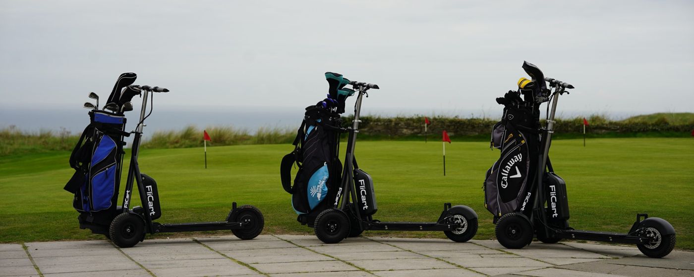 Three FliCart electric golf buggies, each carrying a golf bag, lined up beside a green by the sea